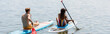 back view of young redhead man and sportive african american woman in striped swimsuit sitting on sup boards and sailing on lake on summer day, banner