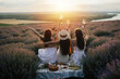Beautiful girlfriends having picnic in the lavender field in summer sunset. They sitting with hands up and enjoying beautiful landscape.