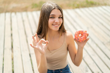 Poster - Teenager girl holding a donut at outdoors showing ok sign with fingers