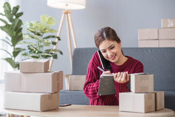 A portrait of a young Asian woman, e-commerce employee sitting in the office full of packages in the background write note of orders use a calculator and smartphone for SME business ecommerce