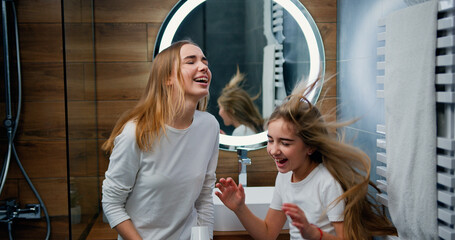 Two funny Caucasian girls with beautiful long hair dancing and singing with hair dryer in modern bathroom at home.