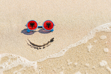 Wall Mural - A painted smile on the sand and sunglasses with the flag of the Albania. The concept of a positive and successful holiday in the resort of the Albania.