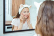 Beautiful little teenage girl with funny cat headband is smiling while looking in bathroom mirror. Portrait of cute little child enjoying skin care procedures