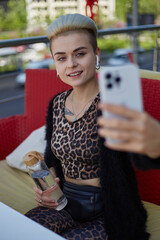 Wall Mural - Cheerful diverse person with short dyed hair taking a selfie photo with a glass bottle of water in hand. Beautiful white woman in leopard print clothes posing for a selfie in a outdoor cafe