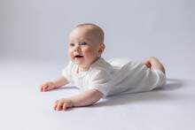 Portrait Of A Cute Baby 5 Months Old In A White Bodysuit On A White Background In The Studio, Smiling Looking Into The Frame. Baby's Health, Newborn Baby, Space For Text