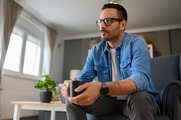 Low angle view of serious young businessman holding coffee cup and contemplating while sitting on armchair. Male freelancer having drink and thinking business ideas while working from home office