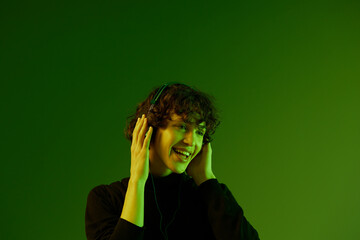 Wall Mural - Man wearing headphones listening to music, dancing and singing with his eyes closed open mouth, DJ happiness and smile, hipster lifestyle, portrait green background mixed neon light, copy space