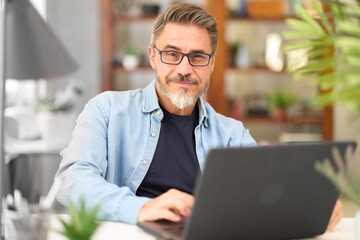 Casual mid adult man with laptop computer at desk in home office, banking online, remote working. Portrait of happy older gray haired bearded guy smiling. Businessman managing business on internet.  