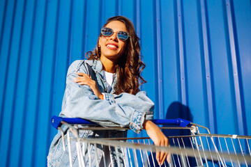 Wall Mural - Young woman with shopping cart having fun near color wall. smilling african woman going shopping. 