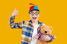 Happy Funny Boy Wearing Thick Glasses And Cap With Propeller. Portrait Of Joyful Teenage Boy In Plaid Shirt Holding Teddy Bear Toy Standing On Yellow Studio Background Showing Thumb Up