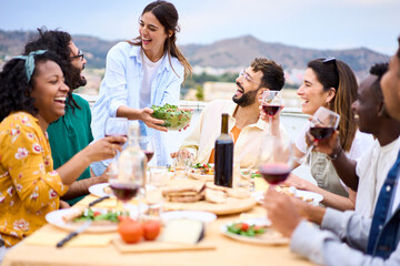 Wall Mural - Young smiling Caucasian woman putting plate with food on table at celebration with happy friends on rooftop. Group of cheerful multiracial friends gathered for barbecue party on outdoor terrace.