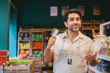 middle-aged latin man owner of an organic grocery store looking at the camera holding a credit card in his hand showing ease of payment. copy space