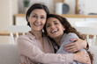© fizkes - Cheerful positive mature 50s mom hugging adult daughter woman with head touch, relaxing on home sofa, looking at camera, smiling, laughing, enjoying closeness, family bonding, motherhood