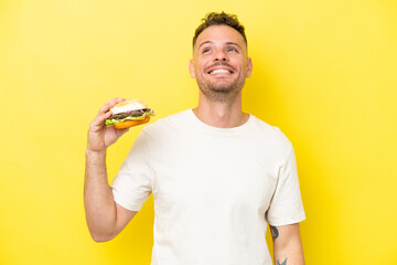 Wall Mural - Young caucasian man holding a burger isolated on yellow background thinking an idea while looking up