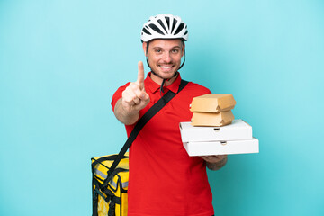 Wall Mural - Young caucasian man with thermal backpack and holding fast food isolated on blue background showing and lifting a finger