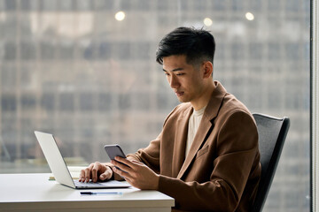 Young busy smart serious Asian Japanese business man executive holding cellphone device using mobile cell phone looking at smartphone working in corporate office with laptop computer technology.