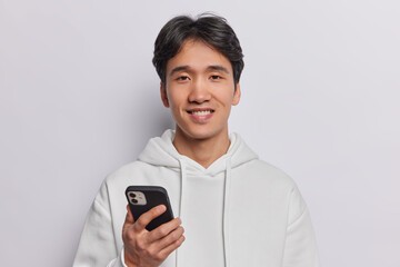 Wall Mural - Horizontal shot of pleased Chinese make teenager uses smartphone sends messages pays for connection to app dressed in casual sweatshirt isolated over white background. Modern lifestyle concept