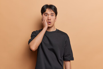 Wall Mural - Surprised shocked dark haired Asian man keeps hand on cheek stares with stupefied expression at camera being terrified by something dressed in black casual t shirt isolated over brown background