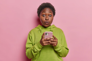 Wall Mural - Studio shot of shocked young Afro woman with short hair holds modern mobile phone dressed in casual green hoodie sends text messages browses internet isolated over pink background. Omg concept