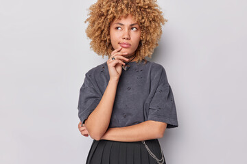 Wall Mural - Curly haired thoughtful woman keeps hand on chin purses lips feels confused dressed in t shirt and pleated skirt poses against white background. People contemplation and making decision concept