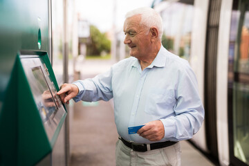 Elderly Caucasian man using cash machine to withdraw his money while standing on tram station.