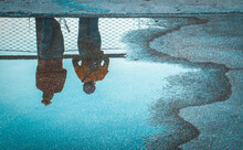 Two People Standing Above Reflection In Water Puddle On Road