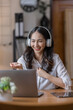 © David - Young adult happy smiling Hispanic Asian student wearing headphones talking on online chat meeting using laptop in home office. College female student learning business education remotely.
