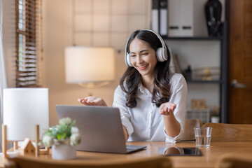 Wall Mural - Young adult happy smiling Hispanic Asian student wearing headphones talking on online chat meeting using laptop in home office. College female student learning business education remotely.