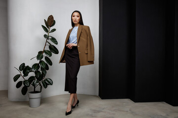 High fashion photo of beautiful elegant young asian woman in pretty brown beige jacket, blazer, blue top, long skirt posing on textured black and white wall, tall flower. Slim figure