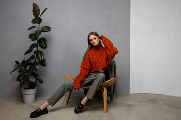 Wall Mural - High fashion photo of beautiful elegant young woman in pretty orange terracotta sweater, gray pants, trousers posing on textured wall, tall flower. Slim figure. Model is sitting on a chair.