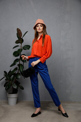 Wall Mural - High fashion photo of a beautiful elegant young woman in a pretty orange shirt, blue pants, trousers, hat, clutch, handbag posing over textured gray wall, tall flower. Slim figure.