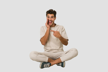 Young caucasian man sitting on the floor isolated on white background saying a gossip, pointing to side reporting something.