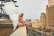 A sexy and charming blonde angel woman in white lingerie with white wings behind her back sits in a seductive pose on the edge of the roof against the backdrop of a cityscape and a gloomy sky.