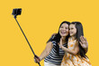 Mom and her daughter are making a common photo on smartphone using selfie stick. Isolated on yellow background.