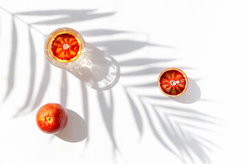 Wall Mural - Creative composition made of half bloody red orange and a glass with cocktail on white background with palm tree leaf shadow. Summer refreshment and party concept. Minimal style.