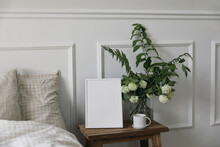 White Picture Frame Mockup And Cup Of Coffee On Wooden Night Stand. White Viburnum, Fern And Solomons Seal Flowers Bouquet. Bedroom View. Beige Pillows, Linen Blanket In Bed. Home Interior Decor.