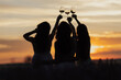 Back view of company of young female friends with hands up having fun, raising glasses with wine and enjoy beautiful sunset at summer picnic in lavender field.	

