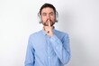 © Roquillo - Surprised businessman wearing blue t-shirt with headphones over white background makes silence gesture, keeps finger over lips and looks mysterious at camera