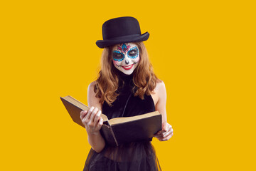 Wall Mural - Happy, smiling, tricky girl in black suit and witch hat with skull makeup reads witchcraft book at Halloween party. Portrait of funny teenager in Halloween costume on an isolated yellow background.