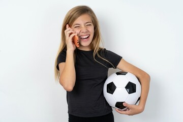 Overemotive happy beautiful caucasian teen girl wearing sportswear and football ball over white wall laughs out positively hears funny story from friend during telephone conversation