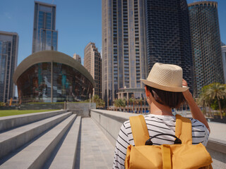 Wall Mural - Enjoying travel in United Arabian Emirates. Young woman with yellow backpack walking on Dubai Downtown in sunny summer day. view from the back or rear view