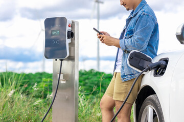 Progressive man with his electric car, EV car recharging energy from charging station on green field with wind turbine as concept of future sustainable energy. Electric vehicle with energy generator.