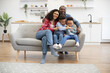 Full length portrait of multiethnic kids with female resting on couch while happy man embracing everyone in kitchen. Playful brothers and merry spouses being involved in relaxing pastime at home.