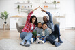 © sofiko14 - Portrait of african american people raising arms high in air forming roof while relaxing on floor of kitchen. Happy spouses making imaginary home while two children staying safe between parents.