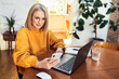 © baranq - Mature woman using mobile phone while sitting in living room with laptop computer