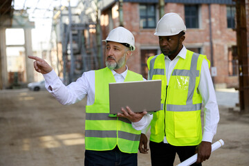 Caucasian elderly builder with wireless laptop explaining all peculiarities of project to african american male architect at construction area. Concept of business meeting and building process.