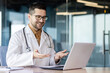 © Tetiana - Successful young doctor inside clinic works online remotely, man consults clients, doctor uses laptop for video call, smiles and consults patient friendly