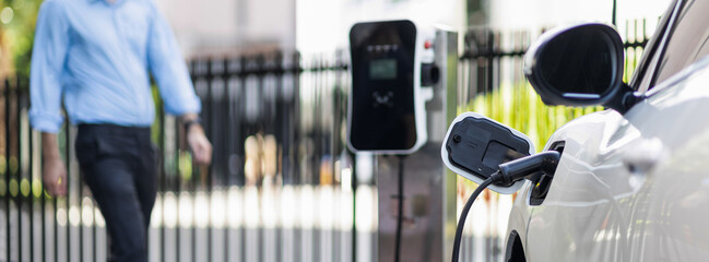 Focus closeup electric vehicle recharge battery at public charging station in the city area with blur businessman in background. EV car attached with electric charger for eco-friendly idea.