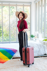 Wall Mural - Asian young happy cheerful female in casual travel outfit standing preparing holding trolley luggage together going on vacation on holiday weekend with rainbow pride LGBTQ freedom flag