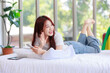 © Bangkok Click Studio - Asian young happy cheerful female LGBTQ lesbian lover laying lying down crossed legs on bed in bedroom smiling using smartphone and tablet computer browsing surfing internet online with rainbow flag